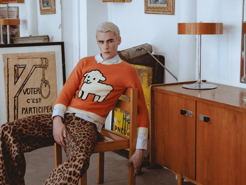 Young man with dyed blonde hair wearing an orange sweater with a sheep graphic and leopard print trousers, seated on a wooden chair.