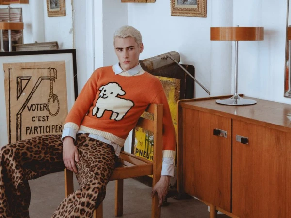 Young man with dyed blonde hair wearing an orange sweater with a sheep graphic and leopard print trousers, seated on a wooden chair.