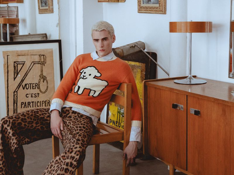 Blonde male model wearing an orange sheep sweater and leopard print trousers seated on a wooden chair in an interior room