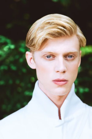 Portrait of a young male model with blonde hair wearing a white high-neck collar against a blurred green foliage background.