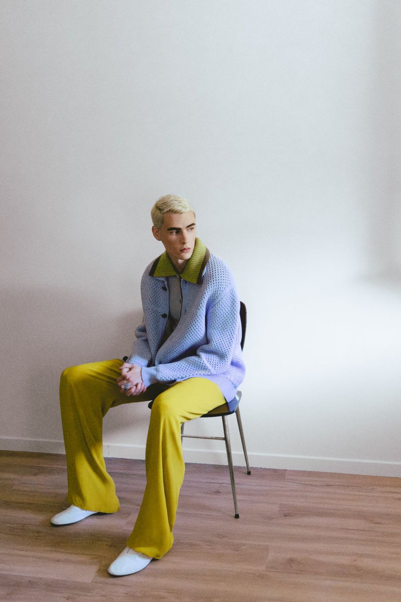 Model with bleached hair seated on a chair wearing a periwinkle knit cardigan, chartreuse trousers, and white shoes against a plain wall.