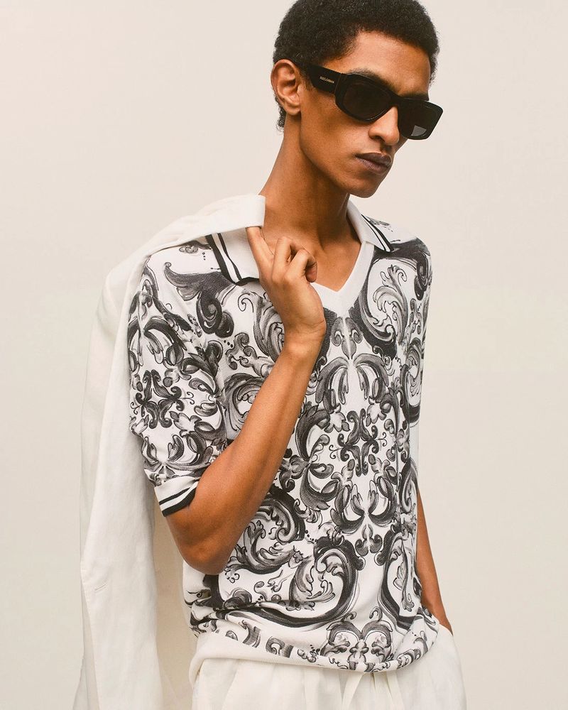 Male fashion model poses in polo shirt with an ornate black and white design. He wears black sunglasses and white pants against neutral background.