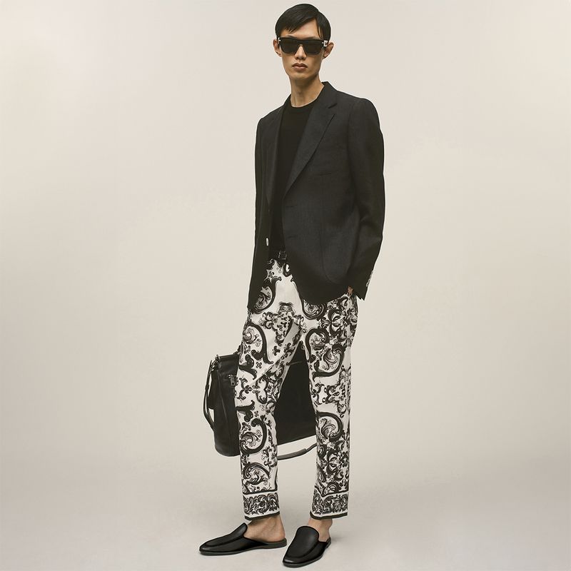 East Asian male model wearing black sunglasses, a black blazer, and black-and-white patterned trousers while holding a black leather bag.