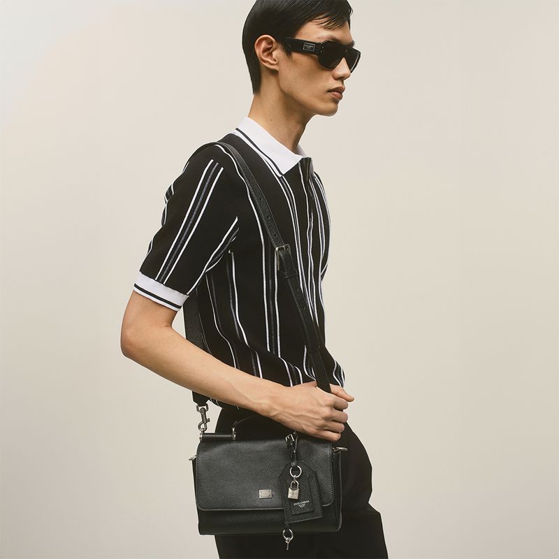 A slim adult male stands indoors with a black striped polo shirt, black sunglasses, and a black leather crossbody bag, against a cream backdrop.