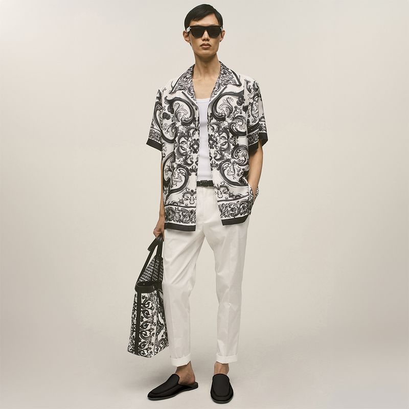 Man in patterned shirt and white pants holds patterned tote bag while posing in a full shot against a plain background.