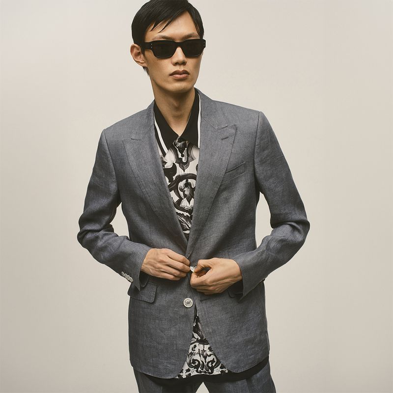 A stylish Asian man in a grey linen suit, wearing sunglasses and a patterned shirt, buttons the jacket in a neutral studio.