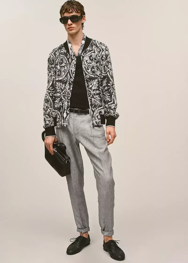 A slender adult male in a patterned jacket, black shirt, grey trousers, and black shoes, holding a briefcase, poses indoors.