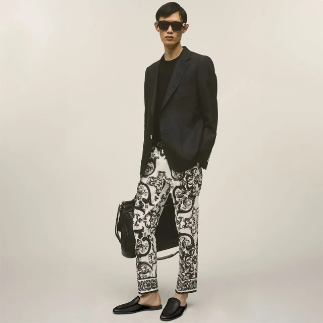 East Asian male model wearing black sunglasses, a black blazer, and black-and-white patterned trousers while holding a black leather bag.