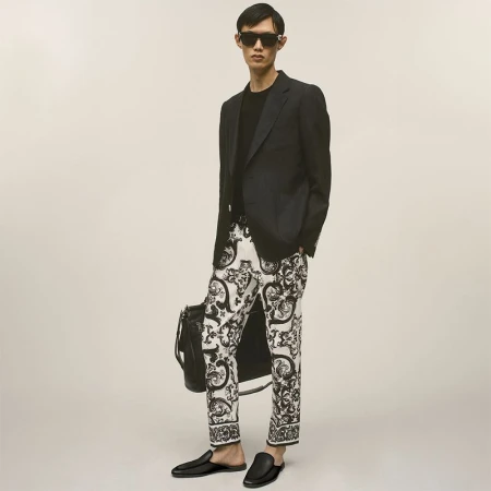 East Asian male model wearing black sunglasses, a black blazer, and black-and-white patterned trousers while holding a black leather bag.