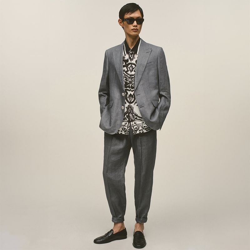 Man wearing a grey suit jacket and trousers with a patterned shirt and dark sunglasses against a seamless neutral backdrop.
