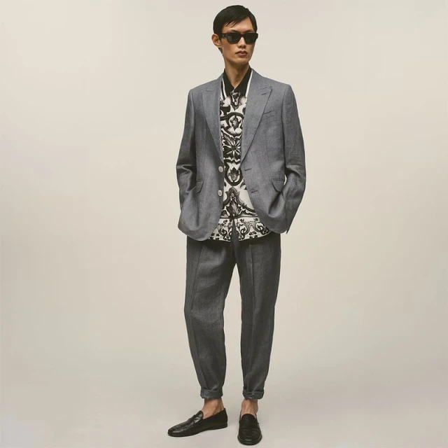Man wearing a grey suit jacket and trousers with a patterned shirt and dark sunglasses against a seamless neutral backdrop.