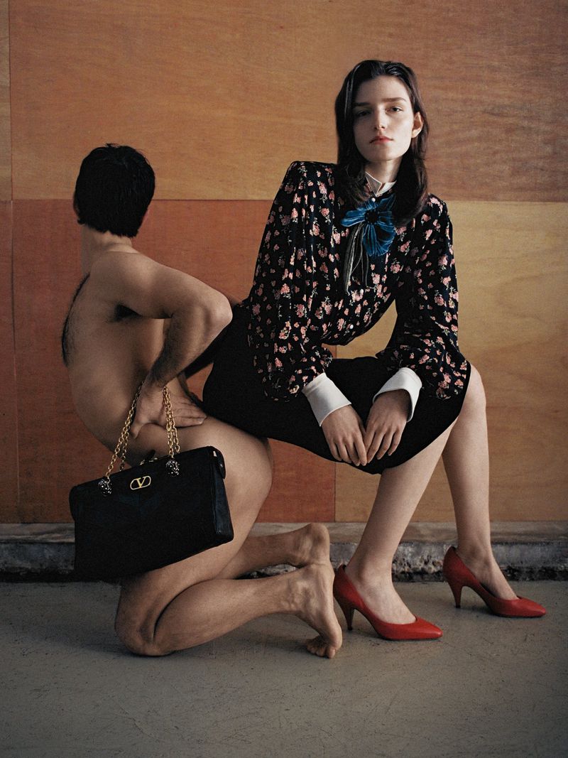 Woman in floral blouse and red heels posing with a kneeling man holding a black handbag against wood paneling