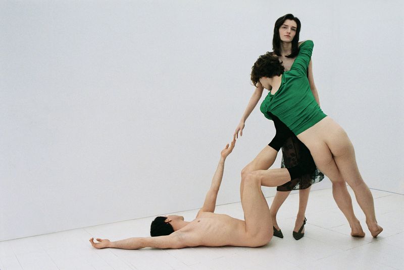 Three figures in a studio setting: one lying down, one standing, and one bending to touch hands. Stark lighting, minimalist backdrop.