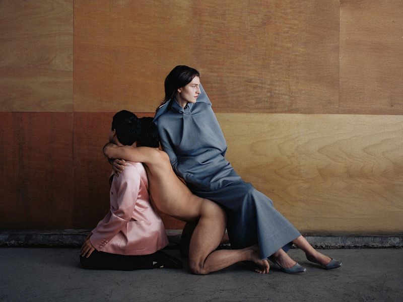 Fashion editorial photograph with a seated woman in a grey dress and two models kneeling in an interconnected pose against a wood panel wall