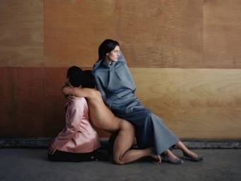Fashion editorial photograph with a seated woman in a grey dress and two models kneeling in an interconnected pose against a wood panel wall