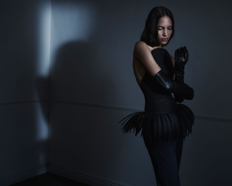 A woman in a black dress and black gloves stands in a dim room, posing with her hands close to her face in an indoor environment.