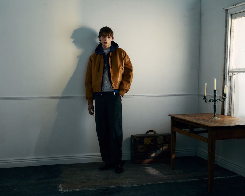 Young male model wearing a brown leather jacket and dark trousers standing against an off-white wall in a dimly lit room.