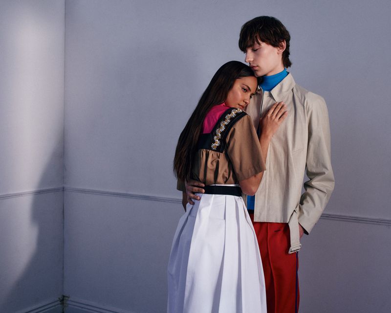Two models embracing indoors against a pale grey wall, wearing layered contemporary apparel.