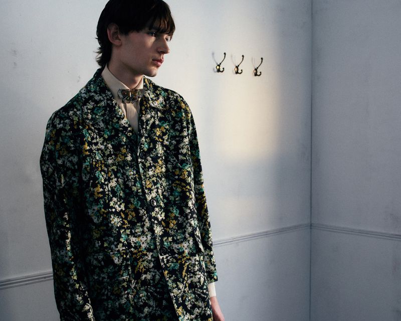 Model wearing a dark floral patterned coat and butterfly collar pin standing indoors against a pale grey wall.