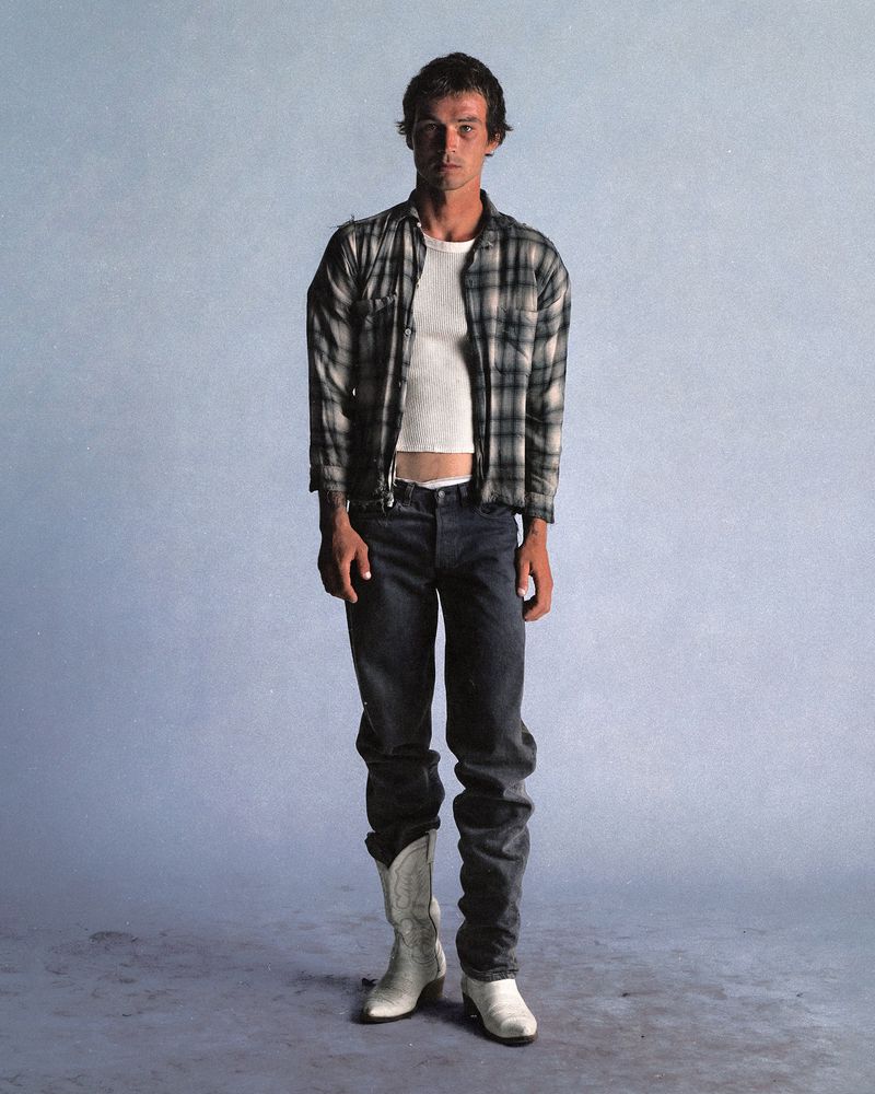 Full-length portrait of a young adult man in plaid shirt, jeans, and cowboy boots, standing in a photo studio with a light blue backdrop.