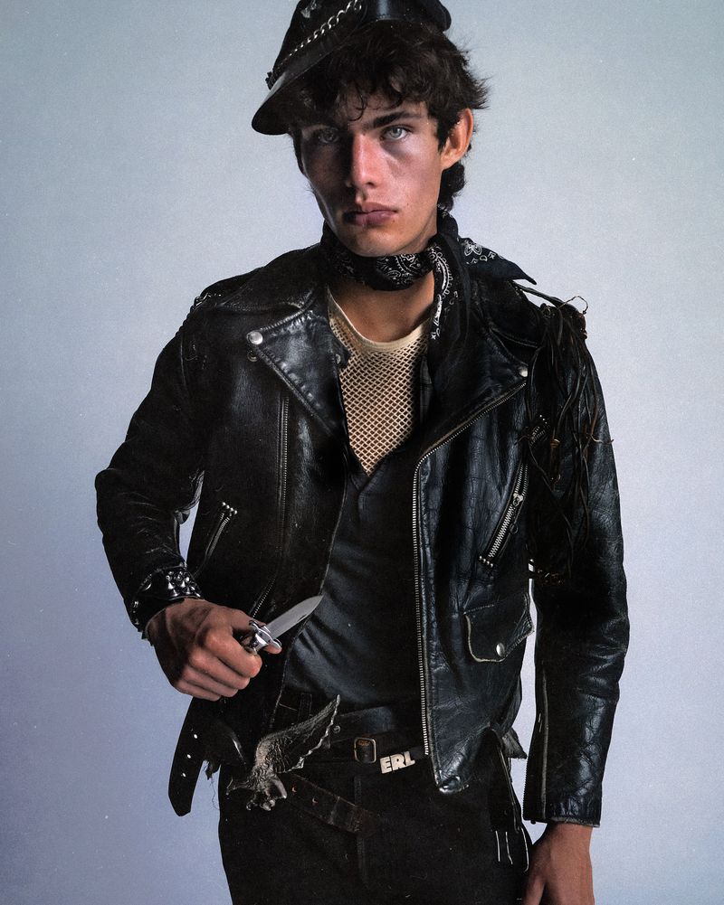 Young man with intense gaze poses in black leather jacket, cap, and bandana, holding a knife, in a studio portrait with a light background.