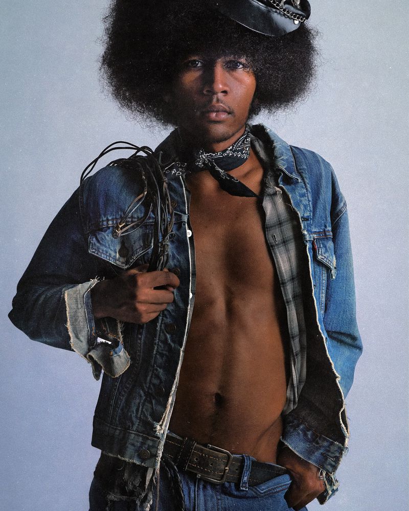 Portrait of a Black adult male with afro, wearing a denim jacket, jeans, and hat, gazing forward against a solid light-grey background.