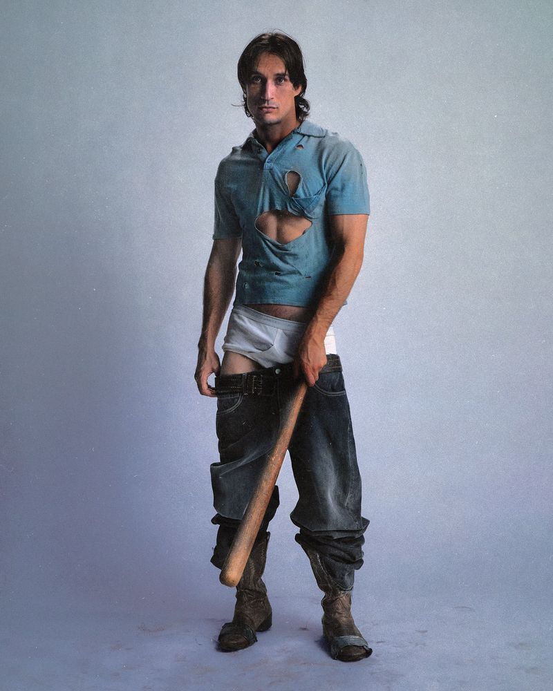 Athletic man with a ripped shirt, loose jeans, and cowboy boots holding a baseball bat posing indoors in a studio-like environment.