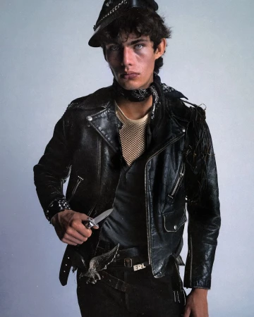 Model in black leather jacket and cap holding a knife against a light grey studio background for ERL Pre-Fall 2025.