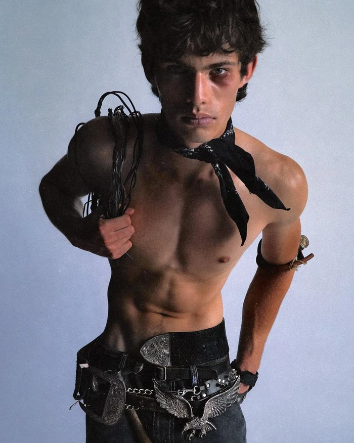 Shirtless male model wearing a black neck bandana and western buckle belts while holding a coiled cord against a studio background.