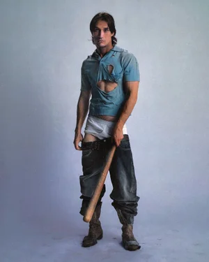 Man wearing a distressed blue polo shirt, white briefs, and sagging jeans holding a wooden baseball bat against a grey seamless background