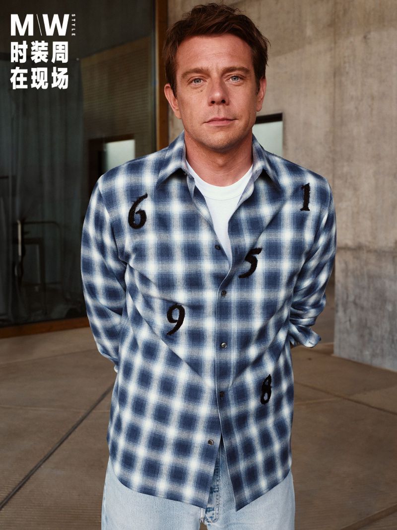 Man wearing a blue and white plaid shirt with black number embroidery against a concrete wall