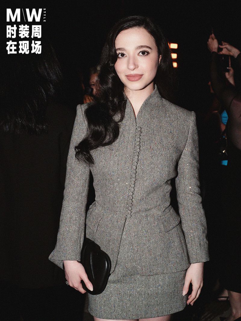 Portrait of Mikey Madison wearing a grey tweed buttoned jacket holding a black clutch in a dark setting