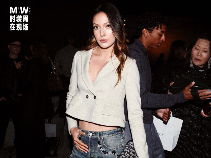 A young woman in a blazer and jeans poses for a fashion event, with event signage visible in the background with other blurred guests.
