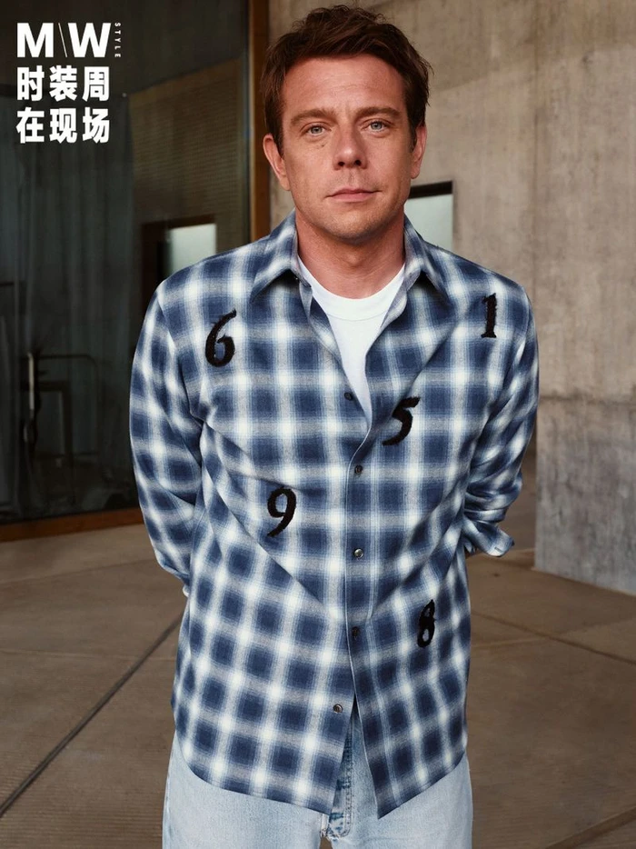 Man wearing a blue and white plaid shirt with black number embroidery against a concrete wall