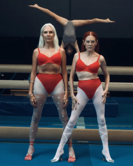 Two adult women in red lingerie and heels pose indoors on a blue floor, with a gymnast upside down in the background.