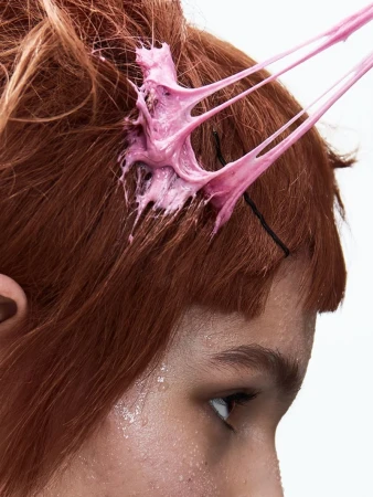 Close-up portrait of a person with red hair and pink bubblegum stuck in strands, shot against a plain white background.