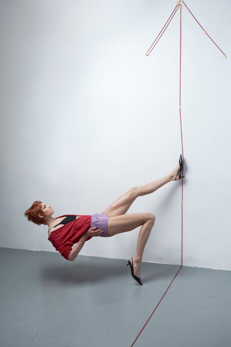 A model wearing a red jacket and purple shorts poses horizontally against a white studio wall with a red string arrow.