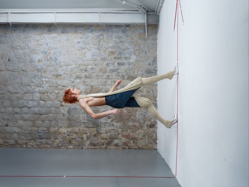 Model with red hair wearing denim jeans and silver high heels pressing against a white wall for The Line of Life project