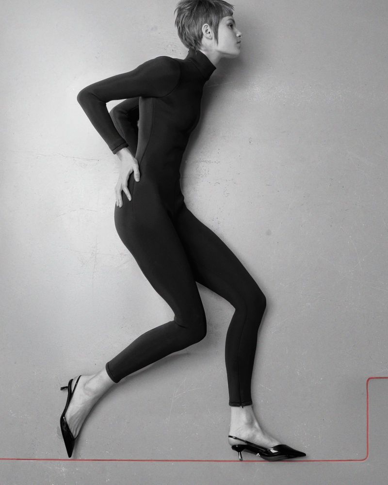 Model with short hair wearing a black catsuit and black slingback heels posing against a grey wall with a red line graphic.