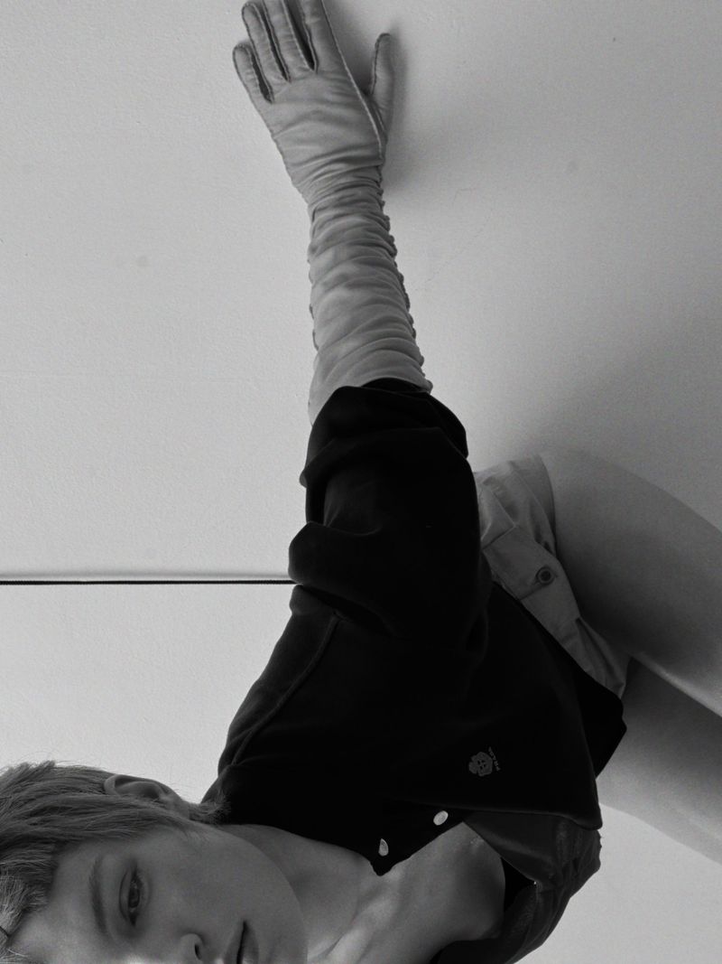 Model with short hair wearing long opera gloves and a dark top reaching toward a plain white wall