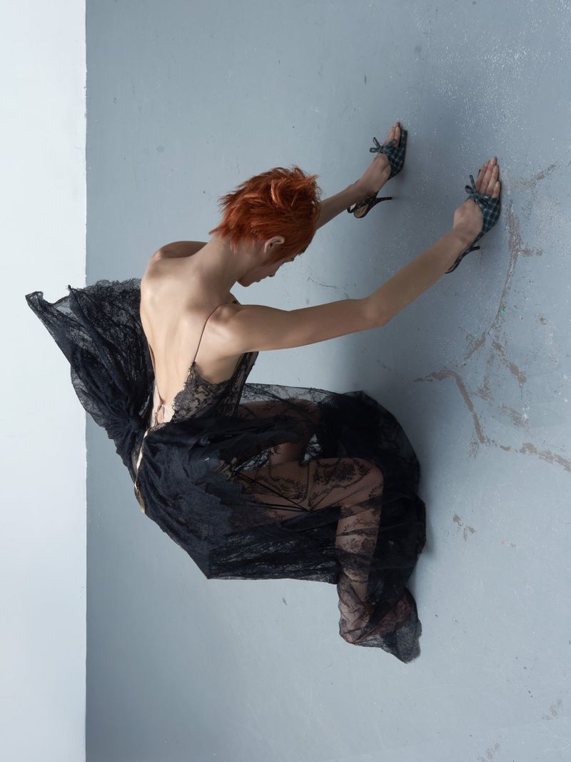 Model with short red hair wearing a black lace dress kneeling on a grey surface for a fashion editorial by Alberto Pelayo.
