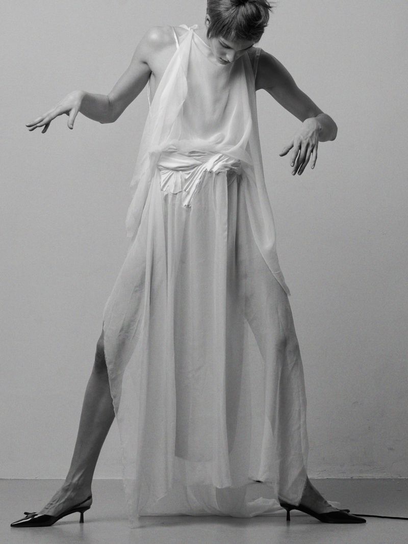 Model in a sheer white layered dress wearing black slingback heels against a seamless grey backdrop.