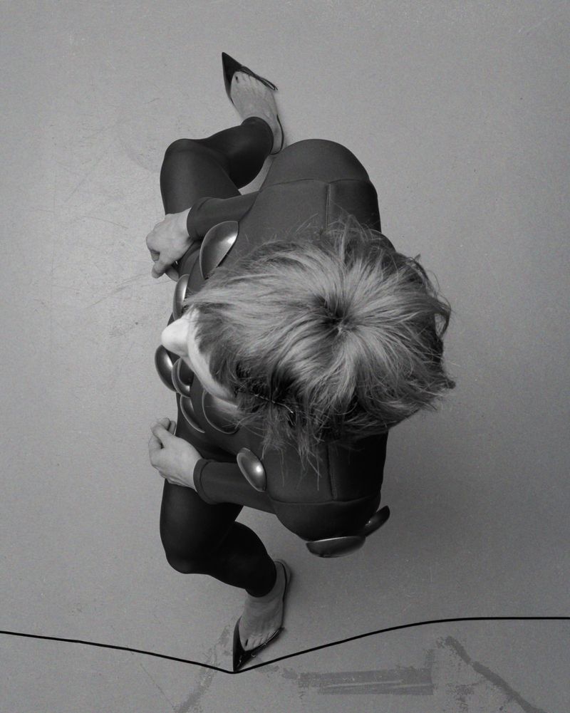 High-angle monochrome fashion photograph of a person wearing a fitted black outfit with rounded accents against a plain surface