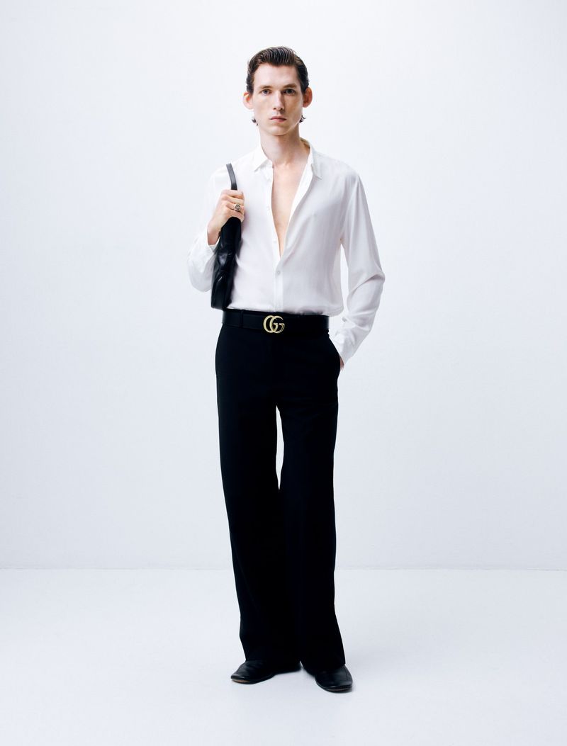 Man wearing a white button-up shirt and black trousers holding a black handbag against a white background.