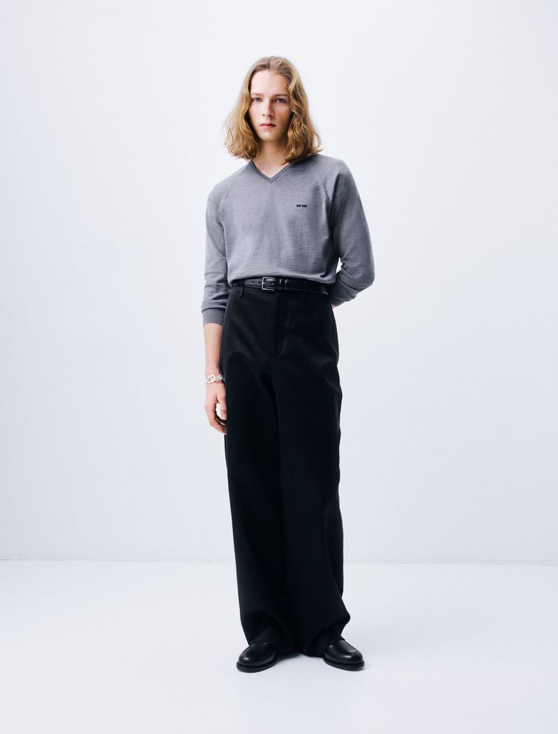 Model with long blonde hair wearing a grey v-neck sweater and black trousers against white seamless background