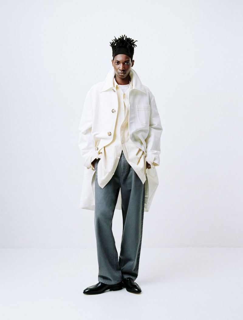 Full-length shot of a Black male fashion model in a white coat, beige shirt, and grey trousers against a white background in a photo studio.