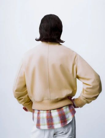 Back view of a person wearing a beige bomber jacket, plaid shirt, and light trousers against a white seamless background