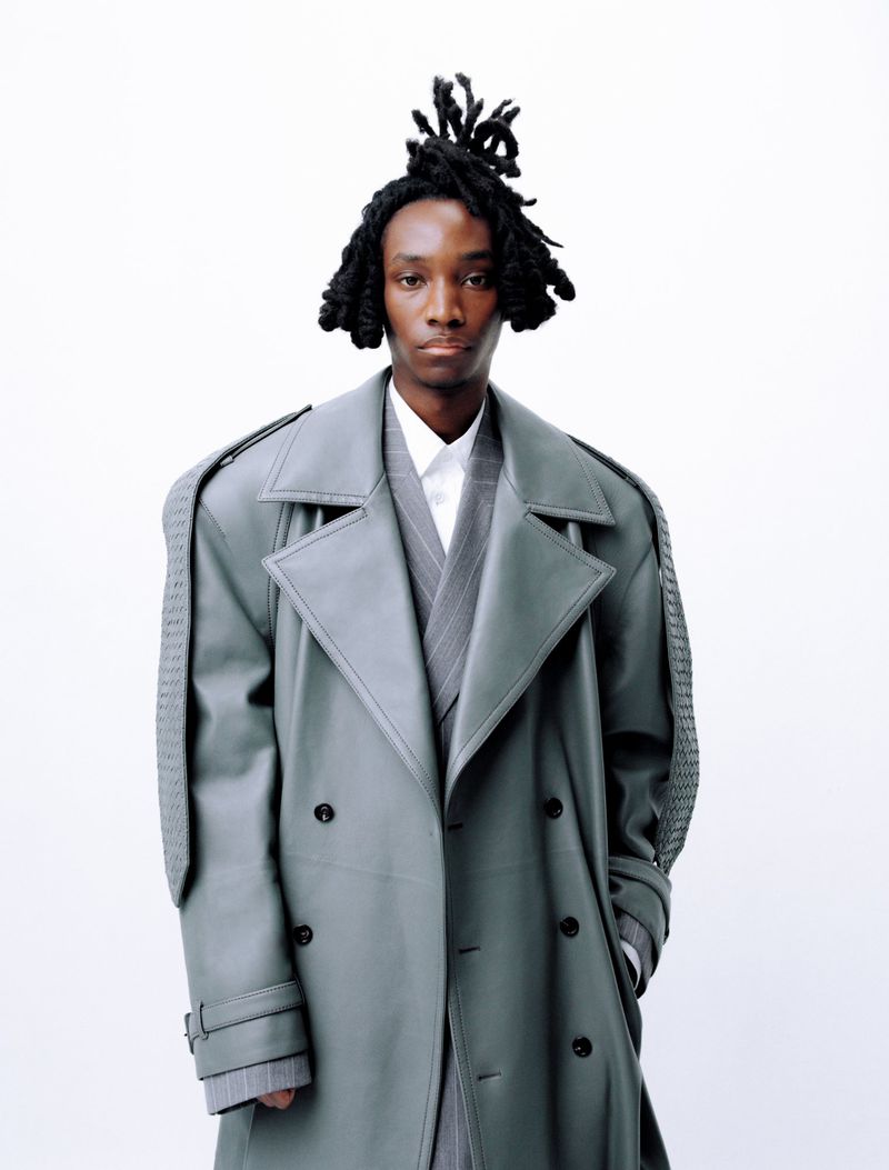 Black man with locs wearing a grey trench coat and pinstripe suit jacket against a seamless white background