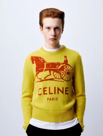 A young male model with red hair wearing a yellow sweater with a printed carriage graphic and red text against a plain white background.