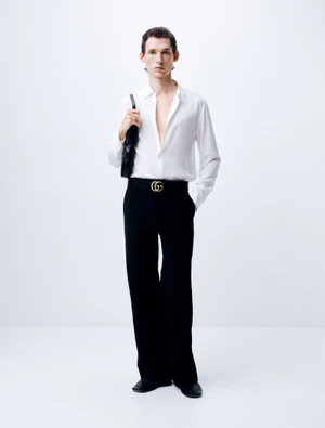 Man wearing a white button-up shirt and black trousers holding a black handbag against a white background.